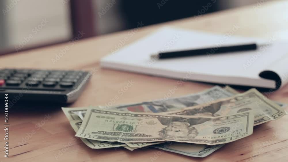 US dollar banknotes on a pile, a calculator and an open paper notebook lie on a wooden table or a desk. Closeup footage, dolly move. US dollar banknotes on a pile, a calculator and an open paper notebook lie on a wooden table or a desk. Closeup footage, dolly move.
