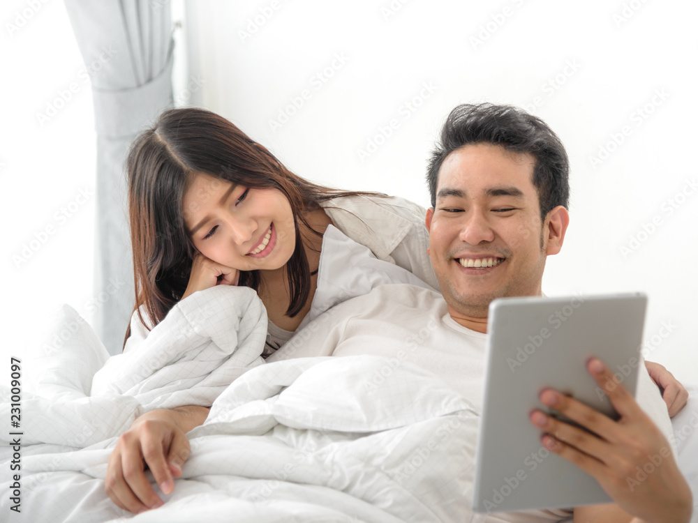 Fototapeta premium Happy Asian couple using laptop and tablet together on bed at home, lifestyle concept.