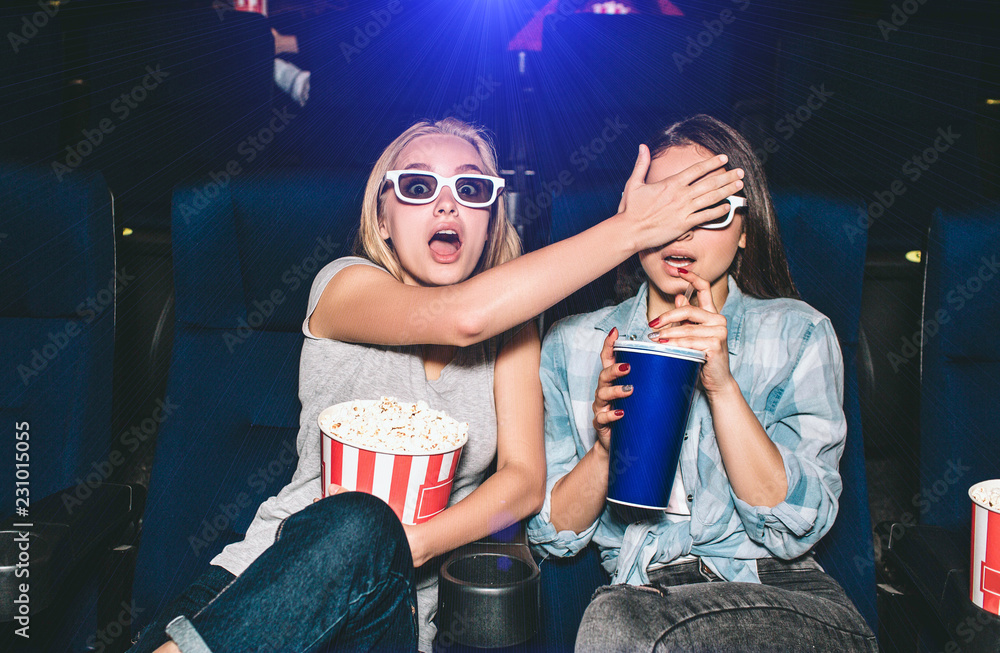 Fototapeta premium Girls are terrified. They are watching a horror movie. Blonde girl is covering eyes of her friend. Brunette is trying to drink some cola from big cup.