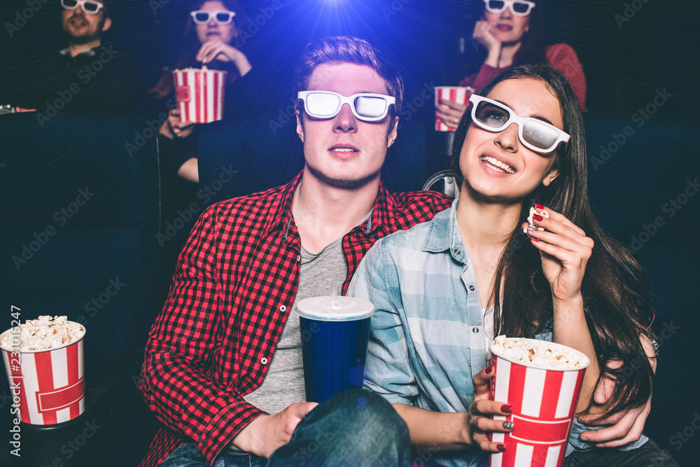 Two young people are sitting close to each other. They have special glasses on their faces to watch movie. Girl is holding basket with popcorn and one piece of it in her hands. Man has a cup of coke. Two young people are sitting close to each other. They have special glasses on their faces to watch movie. Girl is holding basket with popcorn and one piece of it in her hands. Man has a cup of coke.