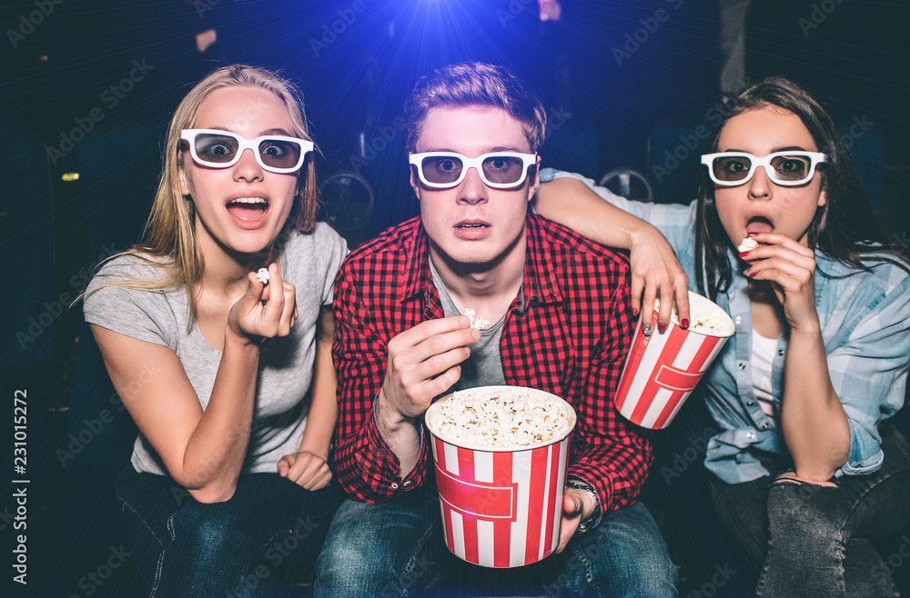 Fototapeta premium Beautiful people in glasses are looking straight forward. They has opened their mouthes. Each of them has a piece of popcorn in their hands. They look excited.