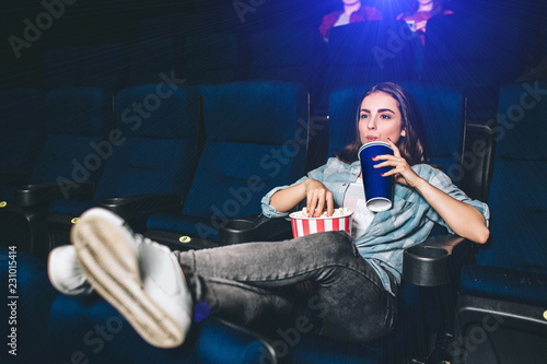 Relaxed and peaceful girl is watching movie. She is keeping her leags in horizontal position towards the floor and drinking coke from blue cup. Also she is taking a piece of popcorn from the basket.