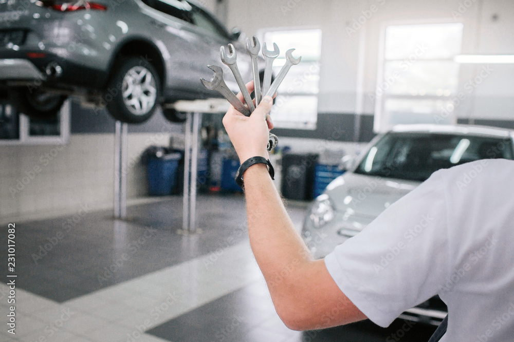 Young and strong man holds set of wrenches in hand. He shows it on camera. Man shows it on camera. He stands in front of cars. Young and strong man holds set of wrenches in hand. He shows it on camera. Man shows it on camera. He stands in front of cars.
