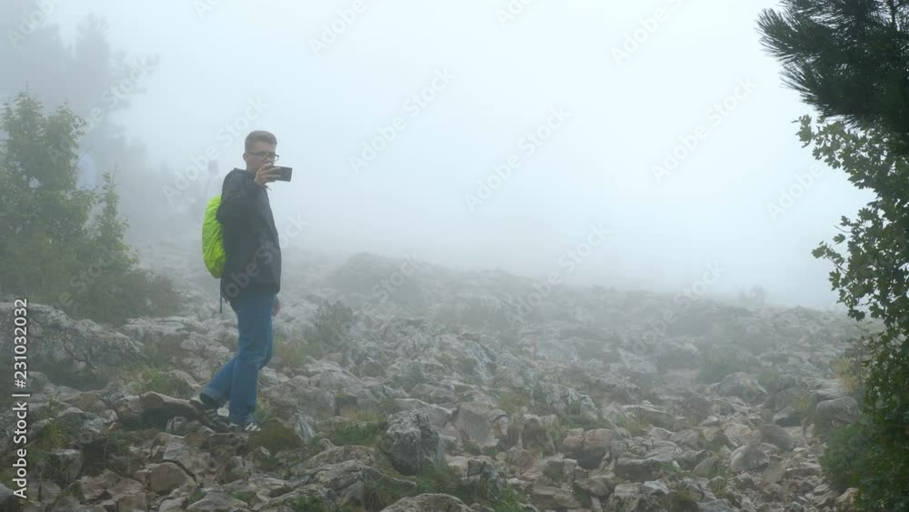 A group of tourists is walking along a gloomy foggy mountain covered with forest. A young man behind the group and taking a photo on a mobile phone. A group of tourists is walking along a gloomy foggy mountain covered with forest. A young man behind the group and taking a photo on a mobile phone.