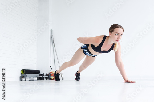 Beautiful young girl in a tracksuit doing exercises for the muscles of the hands and the press in the white gym against the background of sports equipment