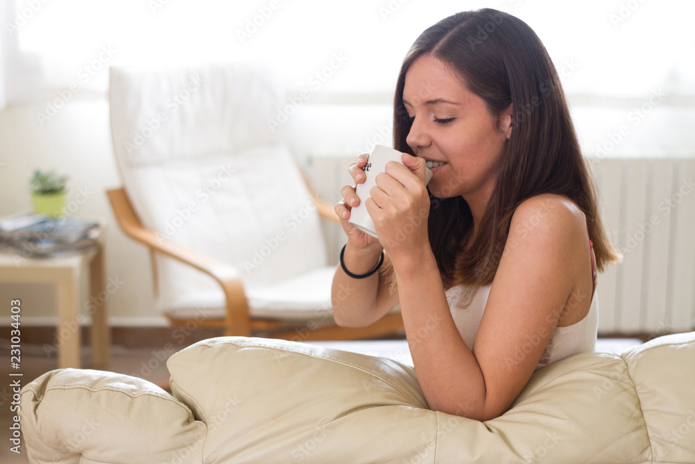Young pretty woman sitting at sofa drinking coffee, tea or infusion and looking outside. Human emotion