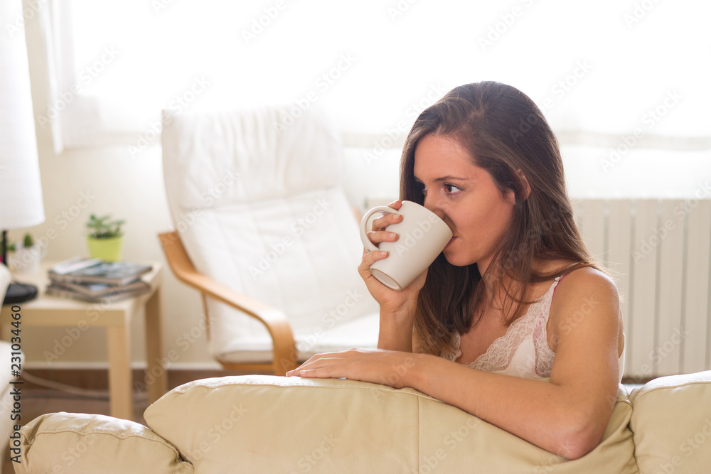 Brunette and young woman looking at a cup of coffee about to take the bowl in an illuminated living room of her house Brunette and young woman looking at a cup of coffee about to take the bowl in an illuminated living room of her house