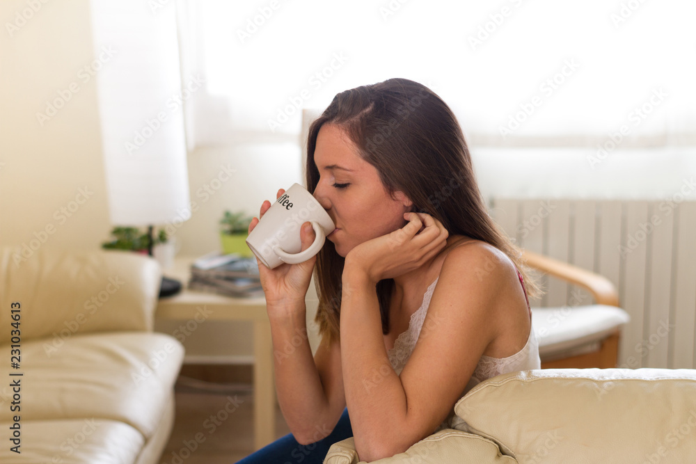Pretty and brunette girl drinking hot drink as coffee or tea sitting on the sofa of her house in the very bright living room Pretty and brunette girl drinking hot drink as coffee or tea sitting on the sofa of her house in the very bright living room