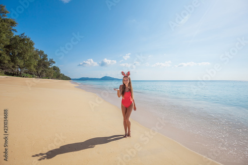 Happy young woman with long hair in red swimsuit wearing funny christmas reindeer antlers holding something imaginary standing on the beach by the sea with blue water and sky on Phuket island,Thailand