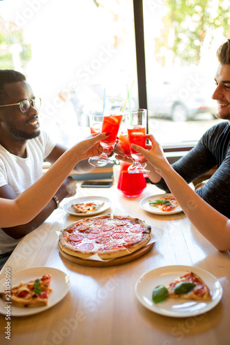 Happy multietnhic friends enjoying appetizer in american bar, close up, cheering with tropical fruits cocktails. Selective focus on female hands holding glasses with pink drinks. photo