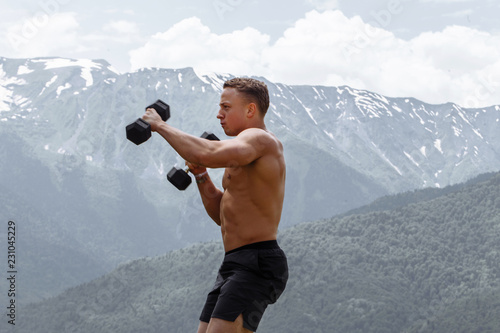 Handsome male boxer with naked torso training shoulder musculs and biceps, doing punches with dumbbells, exercising in open air ober highlands background photo