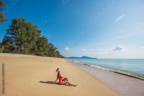 Happy young woman with long hair in red swimsuit and santa claus hat sitting on the wide tropical beach with no people near the sea with beautiful blue water and sky on Phuket island,Thailand