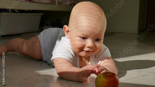 Six-month-old baby lies on the floor and reaches for a red apple. The baby learns to crawl