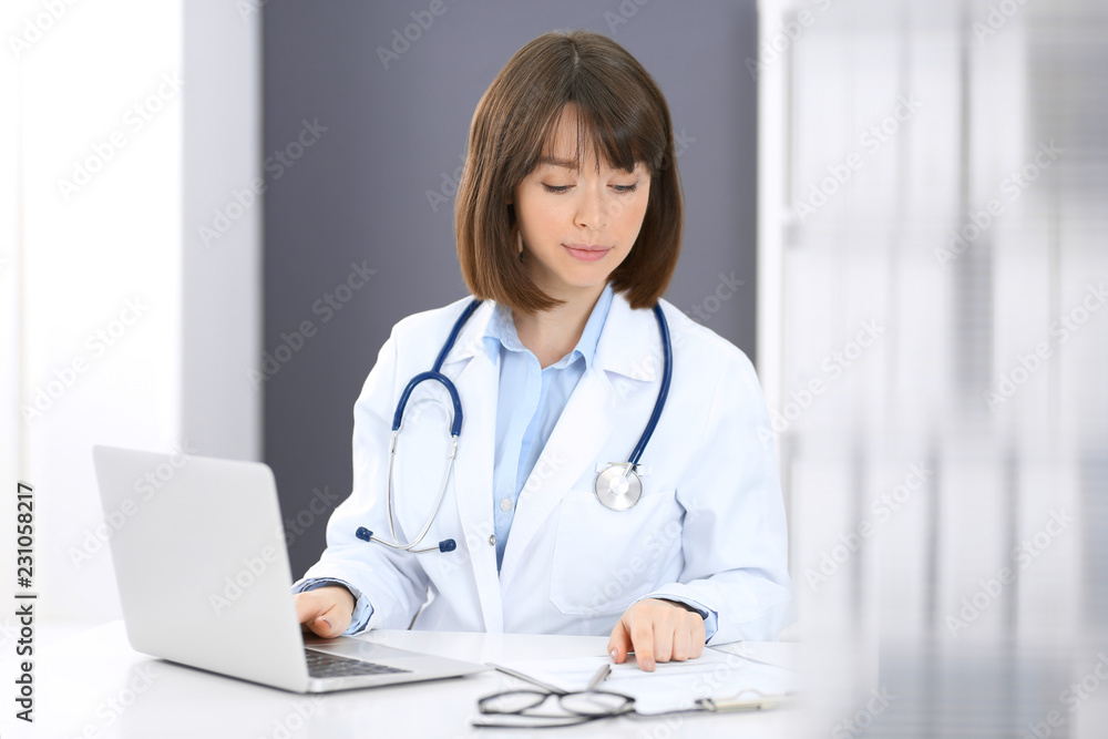 Doctor typing on laptop computer while sitting at the white table in hospital office. Physician at work. Medicine and healthcare concept Doctor typing on laptop computer while sitting at the white table in hospital office. Physician at work. Medicine and healthcare concept
