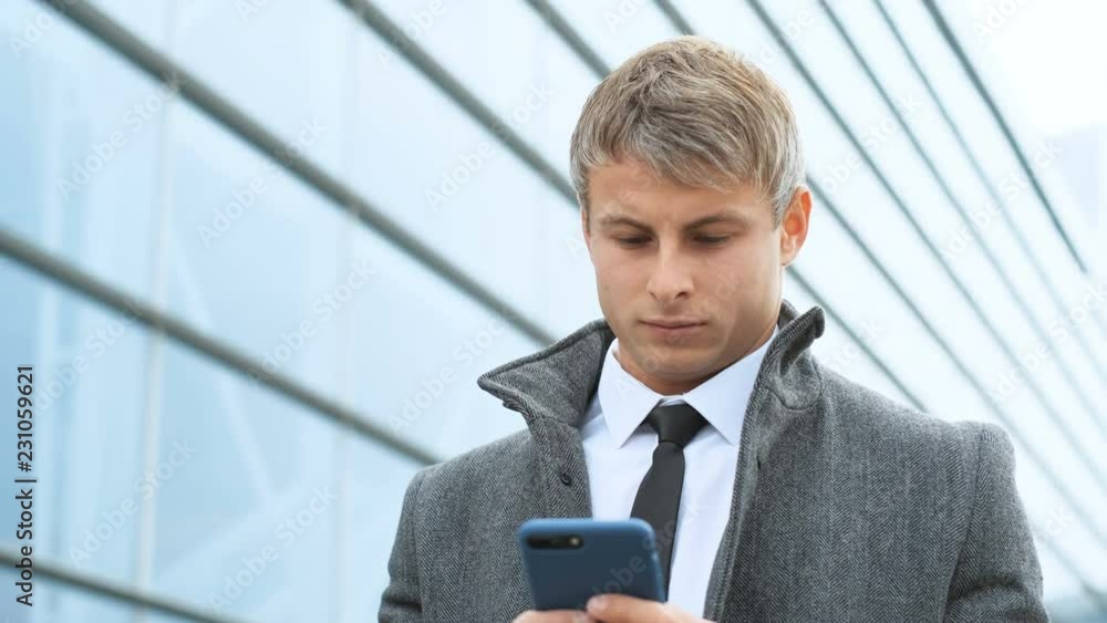 Close up of young businessman in suit holding smart phone, texting, looking at camera, on modern glass building background. Close up of young businessman in suit holding smart phone, texting, looking at camera, on modern glass building background.
