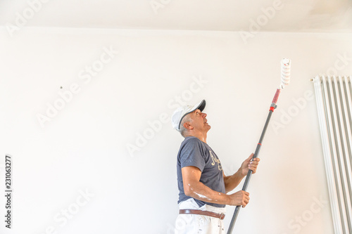 Whitewasher painter man with paint stick roller in hand inside an empty renovation room for painting in white. White wall copy space.