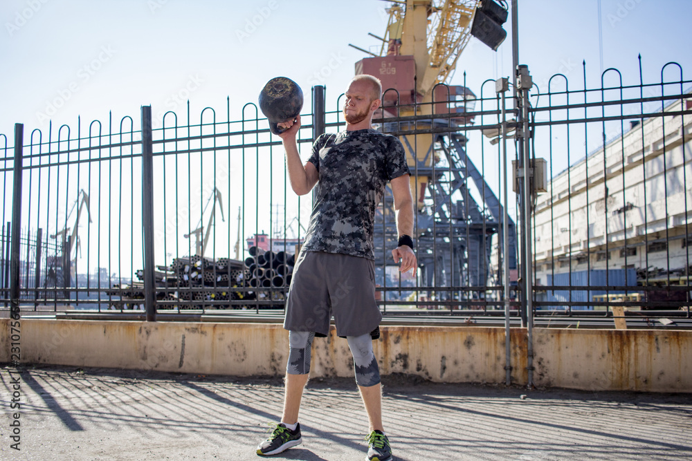 Young bearded male athlete training in industrial zone in sunny day, kettlebells exercises outdoors, urban background - obrazy, fototapety, plakaty 