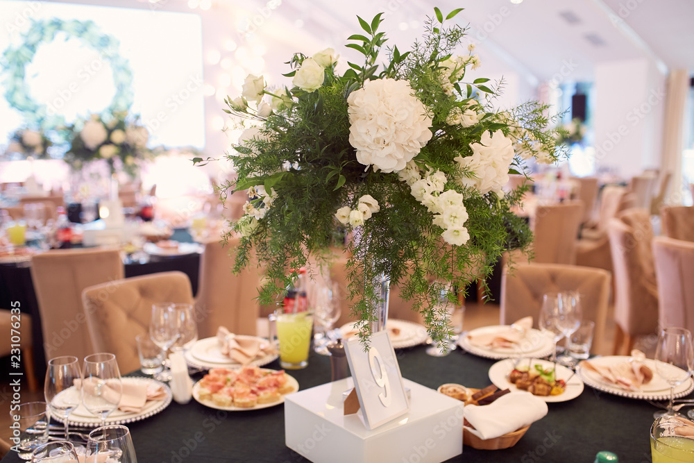 Wedding reception decor. Round dinner tables covered with pink and green clothes and served with glasses, crockery and white flowers stand in the hall