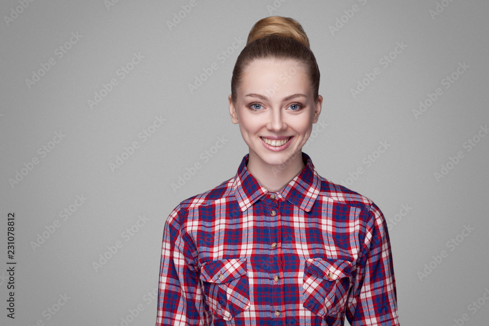 portrait of satisfied beautiful blonde girl in pink checkered shirt collected bun hairstyle and makeup standing and looking at camera with toothy smile. indoor studio shot. isolated on gray background portrait of satisfied beautiful blonde girl in pink checkered shirt collected bun hairstyle and makeup standing and looking at camera with toothy smile. indoor studio shot. isolated on gray background