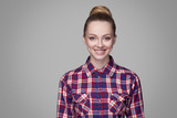 portrait of satisfied beautiful blonde girl in pink checkered shirt collected bun hairstyle and makeup standing and looking at camera with toothy smile. indoor studio shot. isolated on gray background