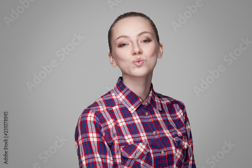Happy kind blonde beautiful girl with pink checkered shirt, collected bun hairstyle and makeup standing and looking at camera and sending kiss. indoor studio shot, isolated on gray background.