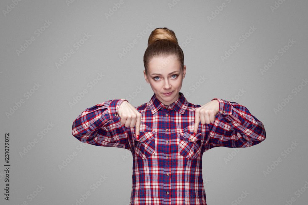 Here and right now. serious angry blonde girl in red, pink checkered shirt, collected bun hairstyle standing and looking at camera and pointing down. indoor studio shot. isolated on gray background Here and right now. serious angry blonde girl in red, pink checkered shirt, collected bun hairstyle standing and looking at camera and pointing down. indoor studio shot. isolated on gray background