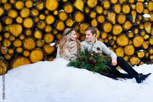 Groom kisses his bride on the temple. Newlyweds with bouquet sits on snow on the wooden background. Winter wedding photo