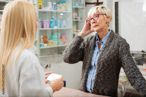Obraz na płótnie Tired, ill mature woman with a headache buying medications in a pharmacy. Holding her head with a hand. Medicine, healthcare and pharmaceutics concept w salonie