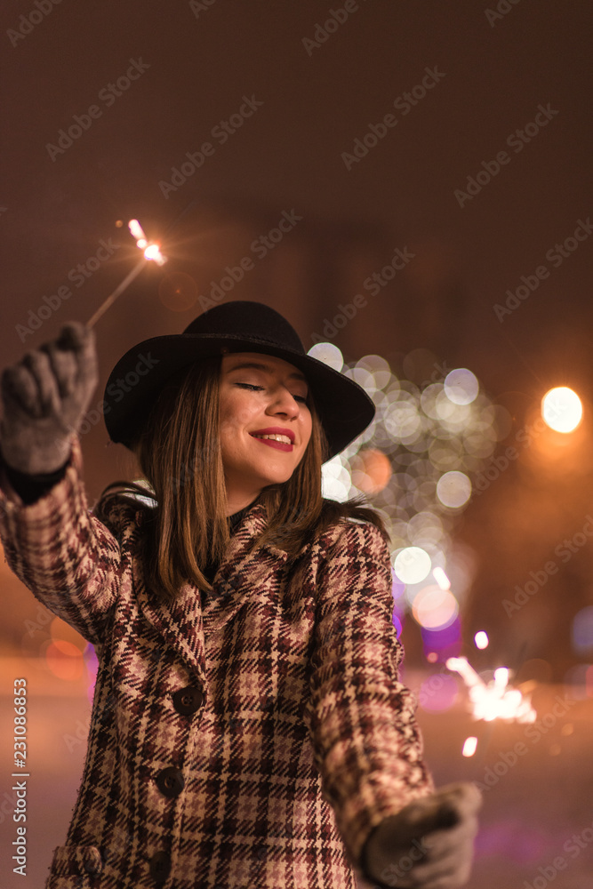 Young girl standing in front on christmas tree lights holding sparklers and smiling at winter alone in the park Young girl standing in front on christmas tree lights holding sparklers and smiling at winter alone in the park