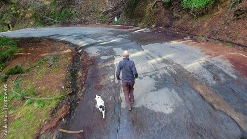 An aerial view following a man trotting down a road with his dog on a leash.