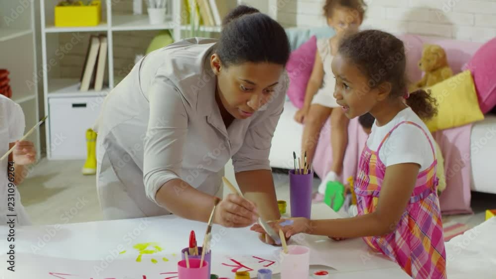 Friendly art teacher using paint brush to apply watercolor on girl’s hand during drawing lesson in art studio or kindergarten Friendly art teacher using paint brush to apply watercolor on girl’s hand during drawing lesson in art studio or kindergarten