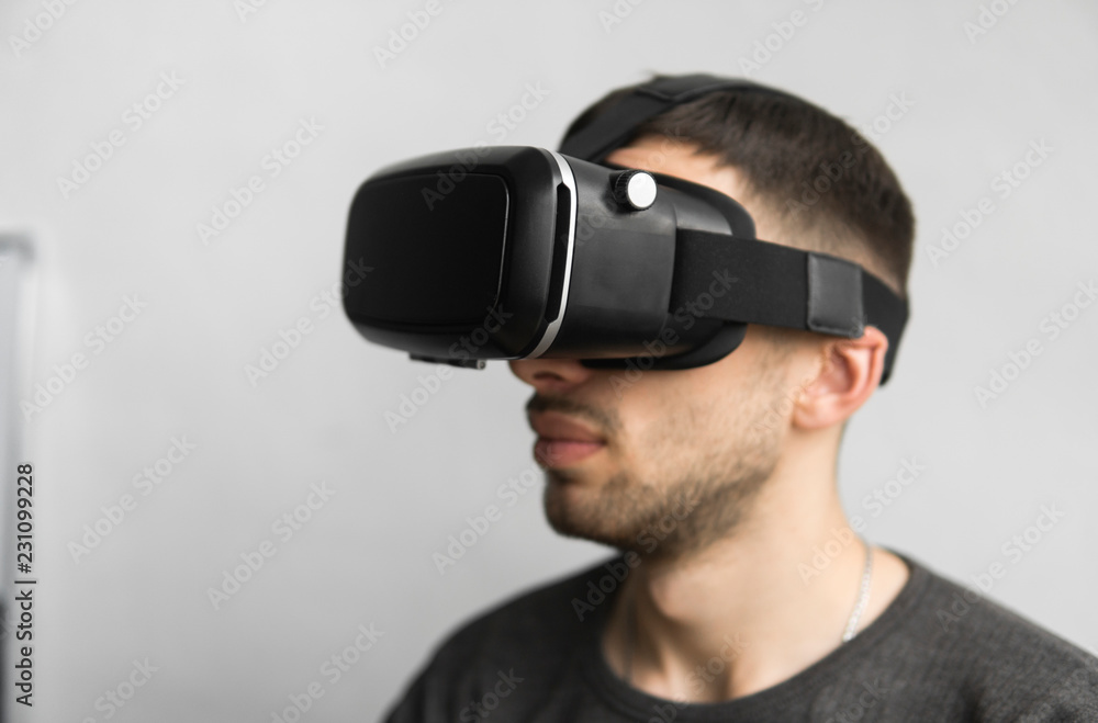 Young man wearing virtual reality goggles headset, vr box and sitting in the office against computer monitor. Connection, technology, new generation, progress concept. Young man wearing virtual reality goggles headset, vr box and sitting in the office against computer monitor. Connection, technology, new generation, progress concept.
