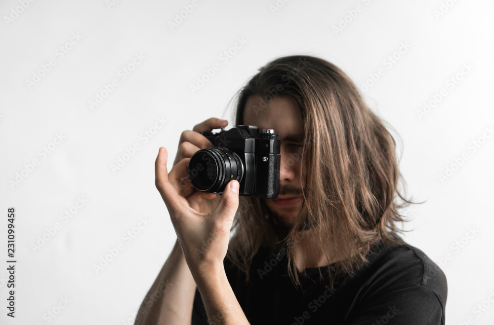 Handsome young bearded man with a long hair and in a black shirt holding vintage old-fashioned film camera on a white background and looking in camera viewfinder. Handsome young bearded man with a long hair and in a black shirt holding vintage old-fashioned film camera on a white background and looking in camera viewfinder.