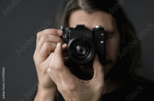 Handsome young bearded man with a long hair and in a black shirt holding vintage old-fashioned film camera on a black background and looking in camera viewfinder. © Volodymyr