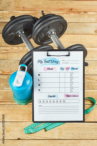 Healthy lifestyle concept. Mock up on workout and fitness dieting diary with copy space. Food diary sheet, measuring tape, blue shaker and dumbbells on a rustic wooden background