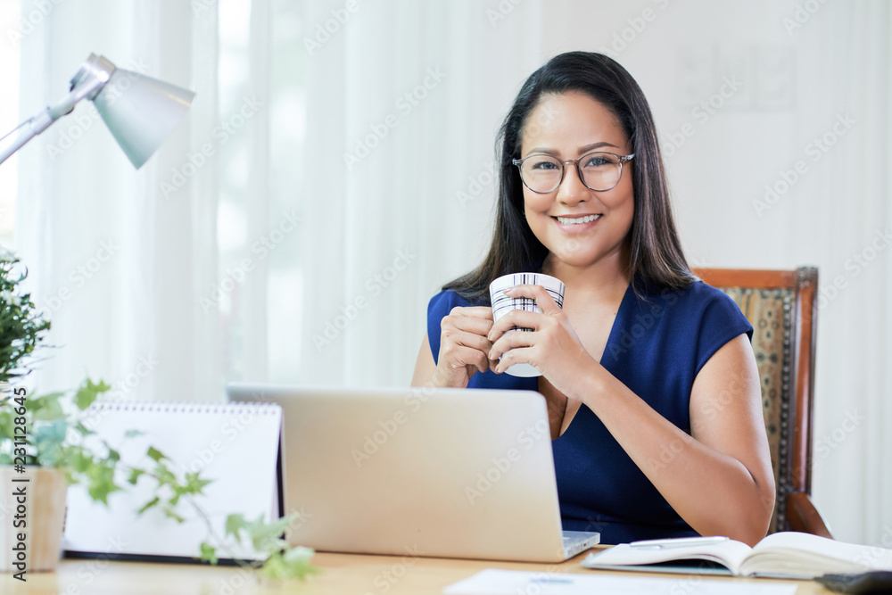 Adult smiling Asian woman in glasses holding cup of coffee sitting at table with laptop in modern office smiling at camera Adult smiling Asian woman in glasses holding cup of coffee sitting at table with laptop in modern office smiling at camera