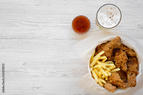 Fast food: fried chicken drumsticks, spicy wings, French fries and chicken fingers in paper box, sauce and cold beer over white wooden background, top view. Flat lay, from above. Copy space. photo