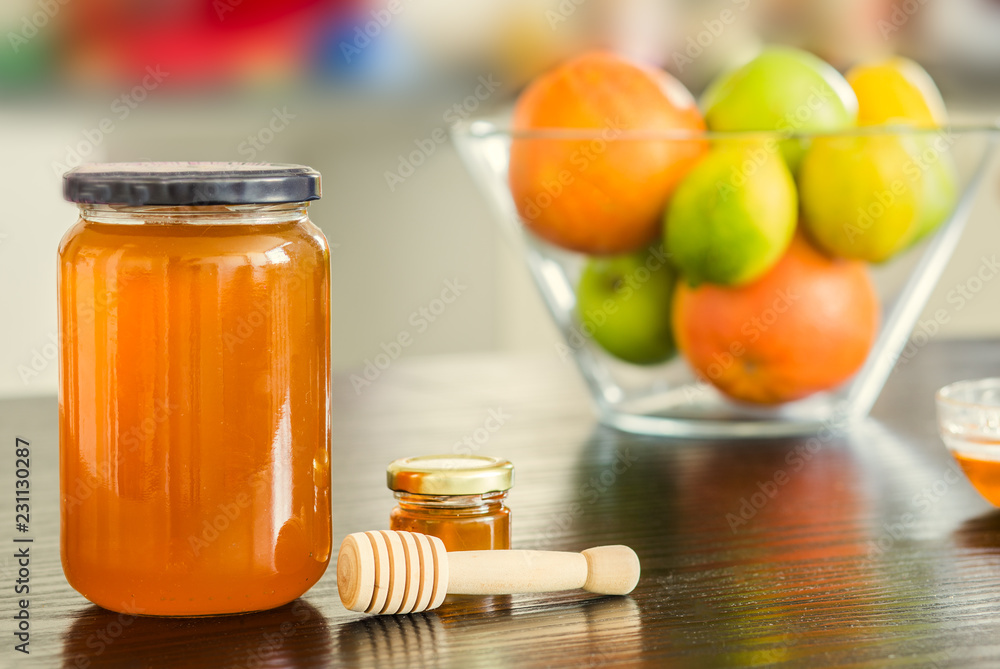 Big and small glass jar of honey with wooden sipper spoon and bowl of fruits on the background. Big and small glass jar of honey with wooden sipper spoon and bowl of fruits on the background.