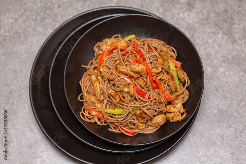 Fried noodles with vegetables, chicken and sesame in a black bowl. Gray background. Close up