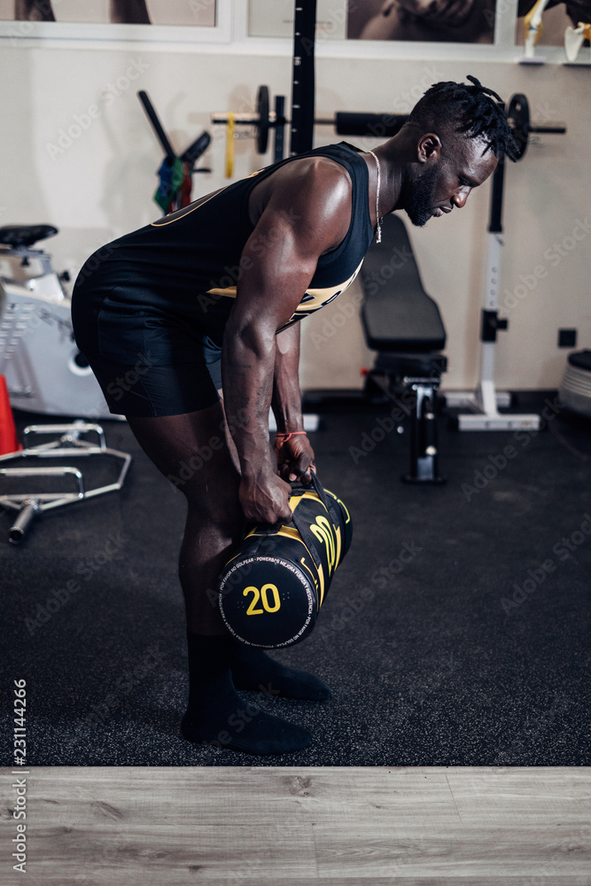 Black sporty young man working out with sandbag while exercises in the gym.  Attractive athletic male doing hard workout with weight training. Sport.  Stock Photo