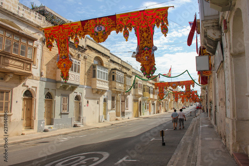 Nearly empty festively decorated street with old traditional balconies. Two adults walking under banners for annual festa religious holiday day in Marsa, Malta