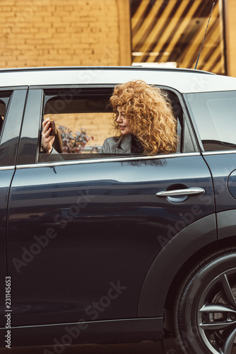 side view of curly redhead woman leaning out from car and taking selfie on smartphone