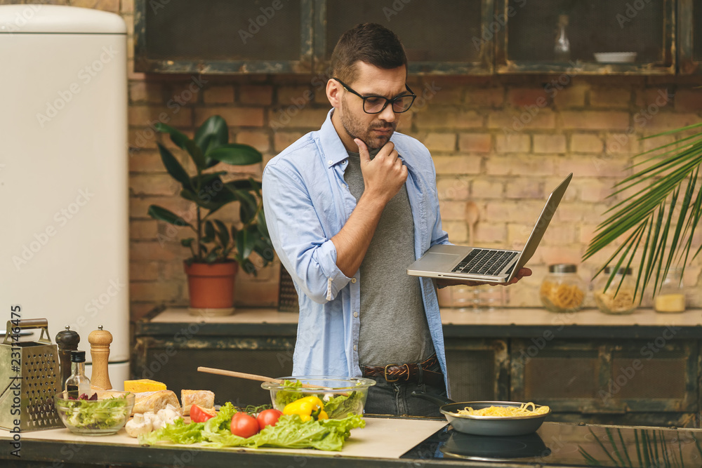 Man preparing delicious and healthy food in the home kitchen on a sunny day. Using laptop computer for searching recipes. Man preparing delicious and healthy food in the home kitchen on a sunny day. Using laptop computer for searching recipes.