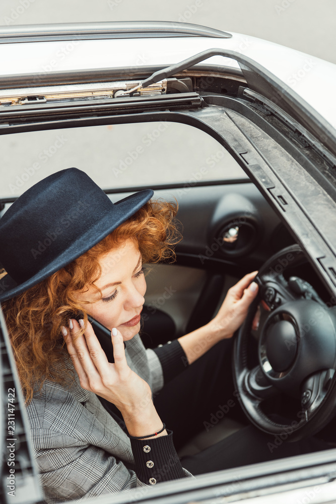 high angle view of redhead woman in black hat talking on smartphone while sitting at steering wheel in car