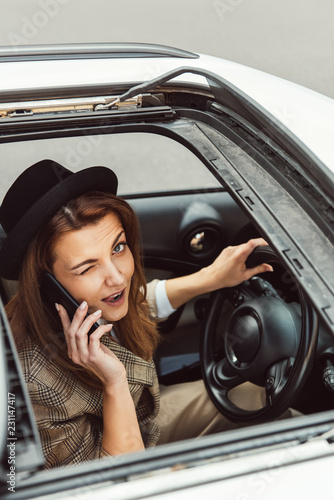 happy woman in black hat winking and talking on smartphone while sitting at steering wheel in car