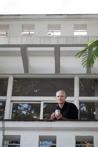 Low angle view of attractive middle-aged Caucasian businessman standing on terrace with cup of tea and smiling at camera