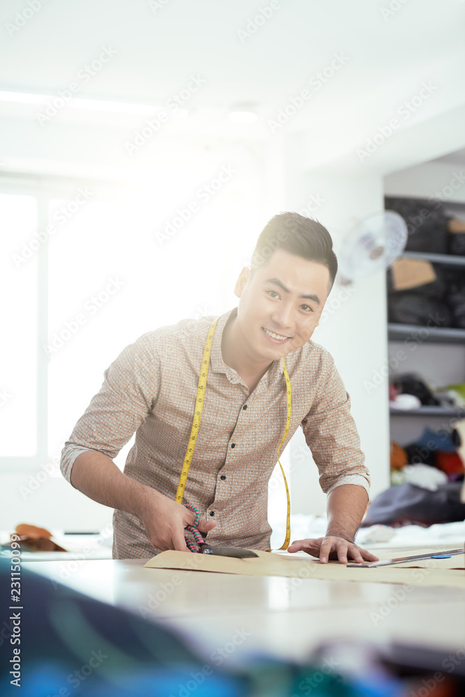 Young male tailor making pattern from fabric while standing at his workplace at atelier