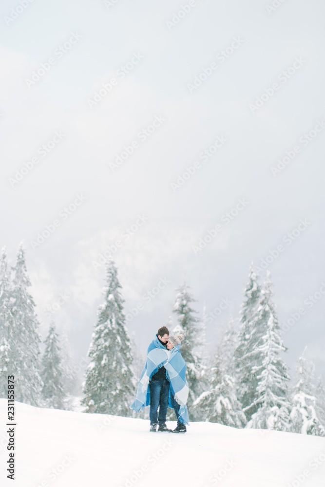 Young handsome man hugs his pretty young girl and covers her with the wool chekered blue blanket. Winter walk in snowy forest. Love in winter