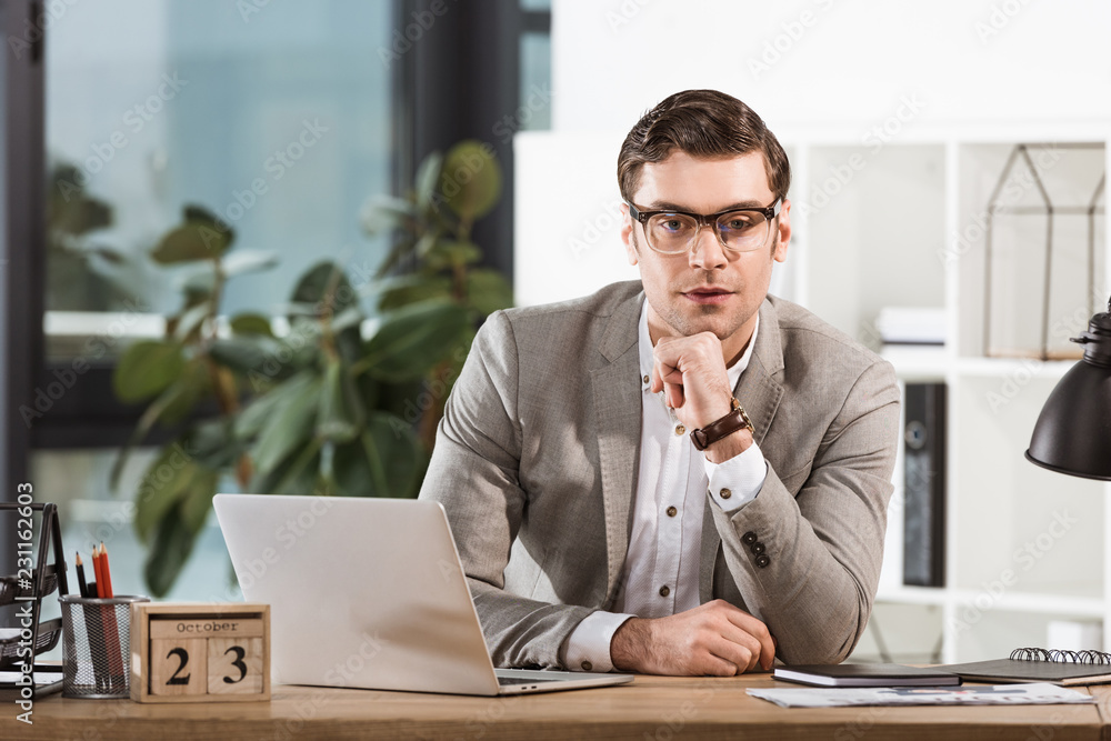 Naklejka premium handsome confident businessman sitting at workplace and looking at camera in modern office