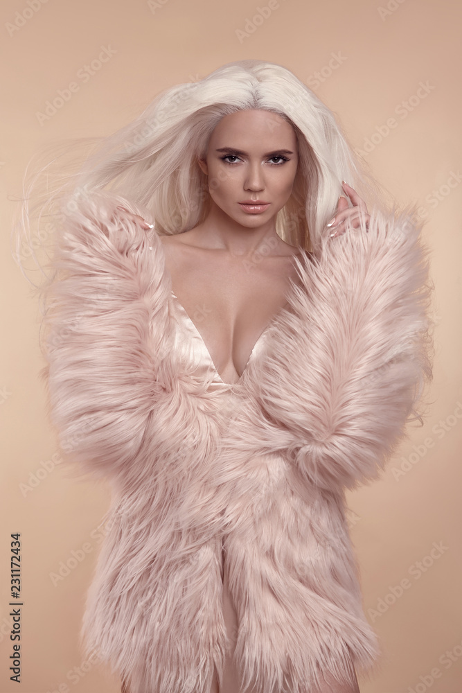 Foto Stock Beautiful blonde stylish woman in fashion winter clothes.  Fashionable sexy girl wearing pink fur coat isolated on beige studio  background. Blond Female with blowing healthy long curly hair. | Adobe